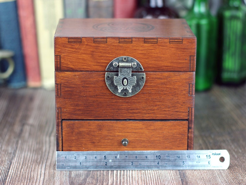 May include: A wooden box with a drawer and a hinged lid, featuring a decorative metal clasp. The box is stained a warm brown color. The box measures approximately 6 inches wide.