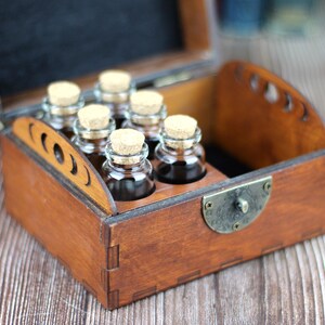May include: A wooden box with a hinged lid, containing six small glass bottles with cork stoppers. The box is stained a warm brown color and has decorative cutouts on the side. The bottles are filled with a dark liquid.