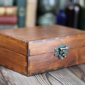 May include: A rectangular wooden box with a hinged lid and a metal clasp. The box is stained a warm brown color, with visible wood grain and dovetail joints. The clasp is an antique brass color.