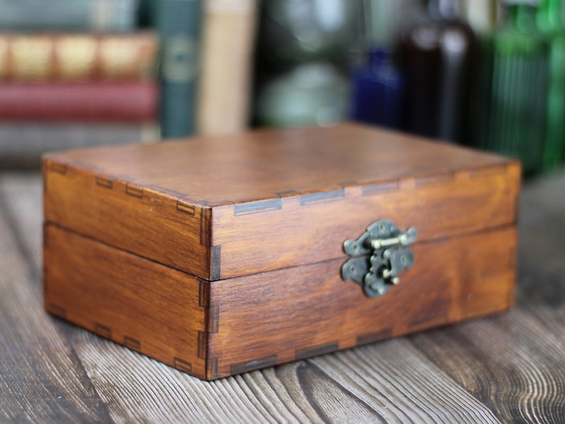 May include: A rectangular wooden box with a hinged lid and a metal clasp. The box is stained a warm brown color, with visible wood grain and dovetail joints. The clasp is an antique brass color.