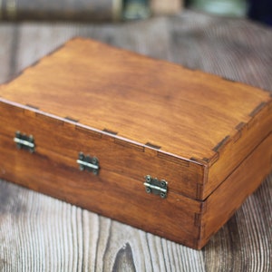 May include: A rectangular wooden box with a warm brown finish. The box features a hinged lid and is secured with three metal clasps. The corners of the box are precisely joined, showcasing the craftsmanship. The box is likely used for storage or display.