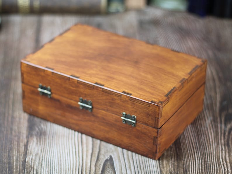 May include: A rectangular wooden box with a warm brown finish. The box features a hinged lid and is secured with three metal clasps. The corners of the box are precisely joined, showcasing the craftsmanship. The box is likely used for storage or display.