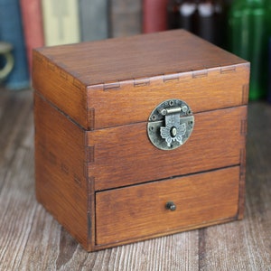 May include: A small, wooden box with a drawer, featuring a dark brown finish. The box has a decorative metal clasp and a small drawer pull. The box is sitting on a wooden surface.