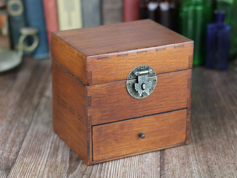 May include: A small, wooden box with a drawer, featuring a dark brown finish. The box has a decorative metal clasp and a small drawer pull. The box is sitting on a wooden surface.