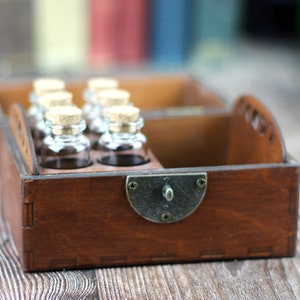 May include: A wooden box with a dark brown finish, holding several small glass bottles with cork stoppers. The box has a metal clasp and a handle on the side. The bottles are arranged in two rows.