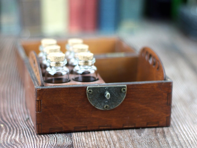 May include: A wooden box with a dark brown finish, holding several small glass bottles with cork stoppers. The box has a metal clasp and a handle on the side. The bottles are arranged in two rows.