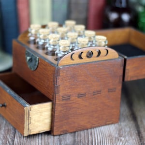 May include: A wooden box with a drawer and a hinged lid, stained a warm brown. The lid features a decorative crescent moon design. Small glass bottles with cork stoppers are visible in the top section. The drawer is open, revealing its interior.