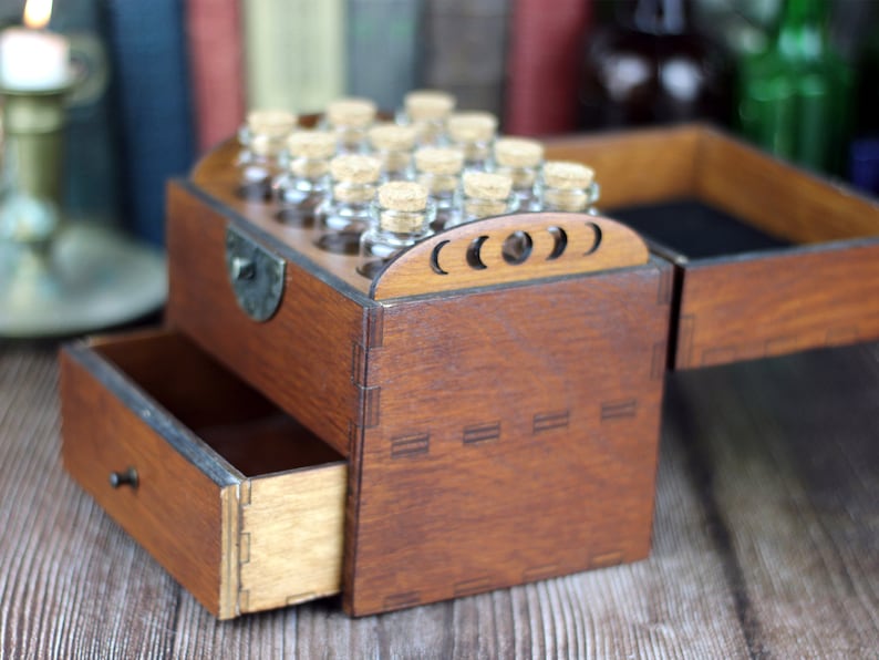 May include: A wooden box with a drawer and a hinged lid, stained a warm brown. The lid features a decorative crescent moon design. Small glass bottles with cork stoppers are visible in the top section. The drawer is open, revealing its interior.