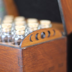 May include: A wooden box with a hinged lid, filled with small glass bottles, each sealed with a cork stopper. The box is stained a warm brown color, and the lid features decorative cutouts. The bottles are arranged neatly inside.