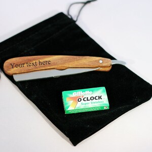 Peut inclure: Un rasoir droit avec un manche en bois et une lame argent&eacute;e repose sur une pochette en velours noir. Le texte "Your text here" est grav&eacute; sur le manche. Une petite bo&icirc;te de lames Gillette 7 O'Clock Super Stainless est &eacute;galement pr&eacute;sente.