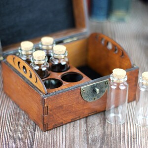 May include: A wooden box with a hinged lid, containing small glass bottles with cork stoppers. Some bottles are filled with a dark liquid. Two empty bottles sit outside the box. The box has a brass clasp.