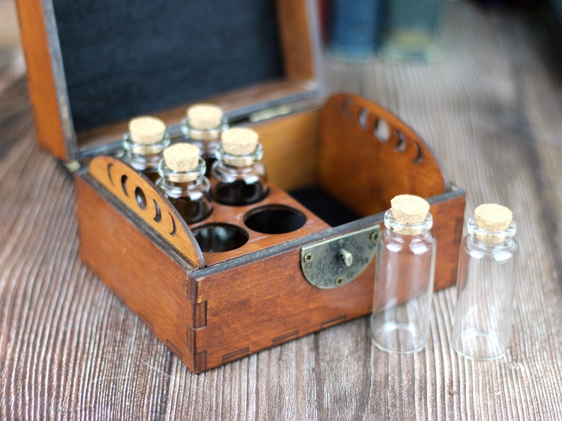 May include: A wooden box with a hinged lid, containing small glass bottles with cork stoppers. Some bottles are filled with a dark liquid. Two empty bottles sit outside the box. The box has a brass clasp.