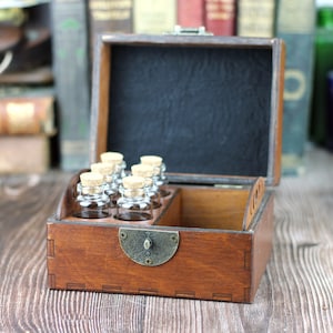 May include: A wooden box with a hinged lid, containing small glass bottles with cork stoppers. The box is stained a warm brown and has a metal clasp. The interior is lined with black fabric. The bottles are arranged in a tray.