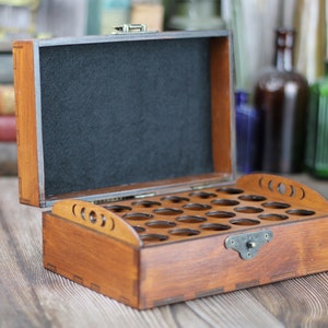 May include: Open wooden box with multiple circular cutouts, likely for holding small items. The box is stained a warm brown color and has a black felt interior. A metal clasp secures the lid. The box is on a wooden surface.