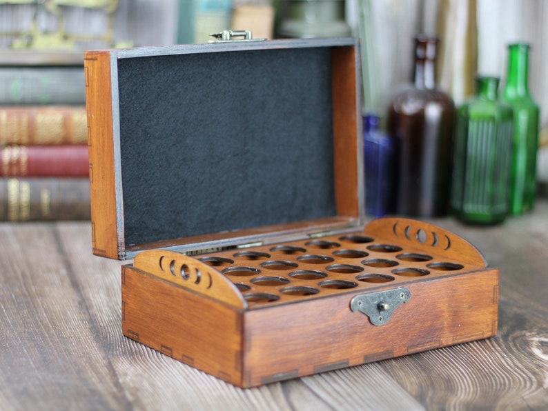May include: Open wooden box with multiple circular cutouts, likely for holding small items. The box is stained a warm brown color and has a black felt interior. A metal clasp secures the lid. The box is on a wooden surface.