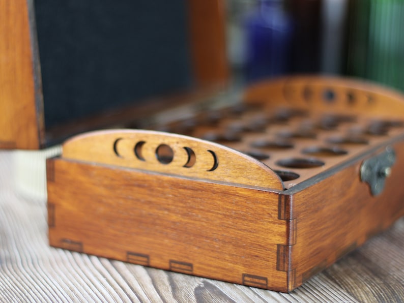 May include: A wooden box with a dark stain, featuring a crescent moon design on the top edge. The box is open, revealing multiple circular compartments inside. A metal clasp secures the lid. The box is likely for storing small items.
