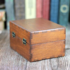 May include: A rectangular wooden box with a hinged lid and metal clasp closures. The box is stained a warm brown color and features visible joinery details. The box is sitting on a wooden surface.