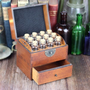 May include: A vintage wooden apothecary chest with a drawer and a hinged top. The top is open, revealing small glass bottles with cork stoppers. The chest is stained a warm brown and sits on a wooden surface. The background includes antique books and bottles.
