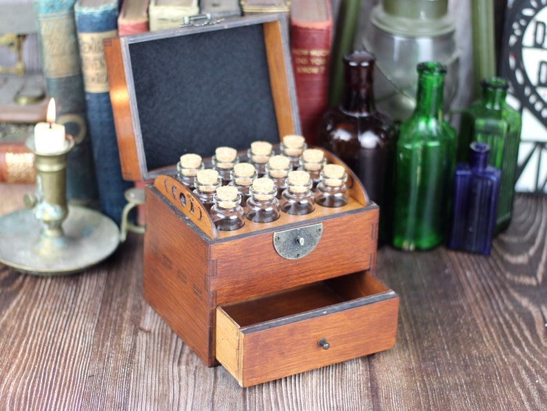 May include: A vintage wooden apothecary chest with a drawer and a hinged top. The top is open, revealing small glass bottles with cork stoppers. The chest is stained a warm brown and sits on a wooden surface. The background includes antique books and bottles.