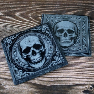 May include: Two square, dark grey slate coasters with engraved skull designs. One coaster features a skull within a floral circle, the other a skull in a decorative frame. The coasters are on a wooden surface.