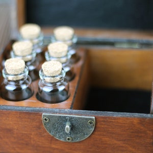 May include: A wooden box with a hinged lid, containing six small glass bottles with cork stoppers. The box has a dark brown finish and a metal clasp. The bottles are arranged in a row, and the box appears to be designed for storage or display.