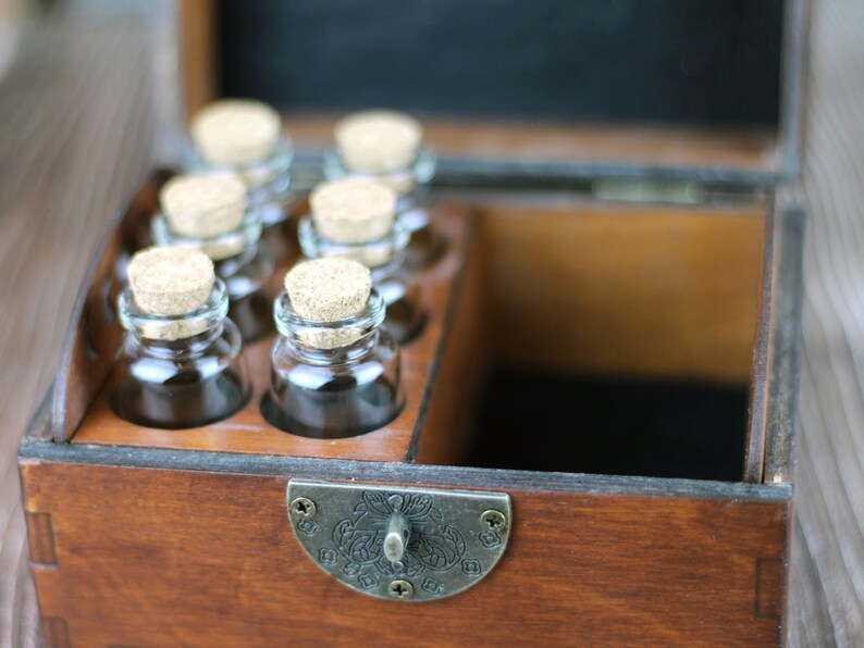 May include: A wooden box with a hinged lid, containing six small glass bottles with cork stoppers. The box has a dark brown finish and a metal clasp. The bottles are arranged in a row, and the box appears to be designed for storage or display.