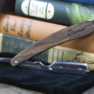 Peut inclure: Un rasoir droit avec un manche en bois, grav&eacute; du nom "Desiree". Le rasoir est ouvert, r&eacute;v&eacute;lant une lame argent&eacute;e. Il repose sur un tissu noir, avec des livres anciens en arri&egrave;re-plan.