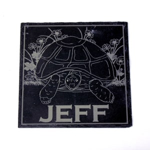 May include: Square black slate coaster featuring an etched turtle design, with flowers and foliage. The name "JEFF" is etched in grey. The coaster is approximately 10 cm square. A decorative item.