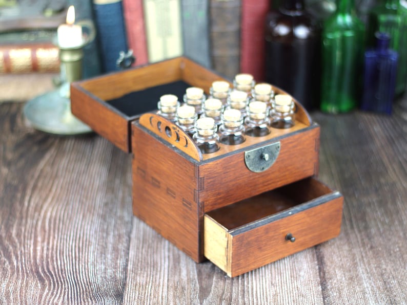 May include: A wooden apothecary chest with a drawer and a hinged lid. The lid is open, revealing small glass bottles with cork stoppers. The chest is stained a warm brown color and has decorative cutouts on the front.