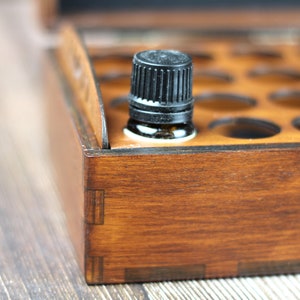 May include: A wooden essential oil storage box with a dark brown finish. A small amber glass bottle with a black cap sits in the foreground. The box has multiple circular cutouts for holding bottles.