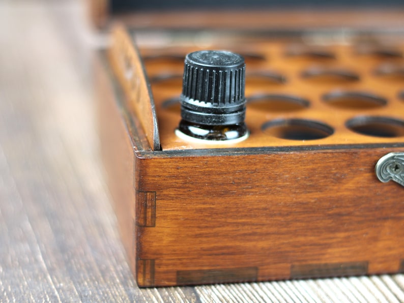 May include: A wooden essential oil storage box with a dark brown finish. A small amber glass bottle with a black cap sits in the foreground. The box has multiple circular cutouts for holding bottles.