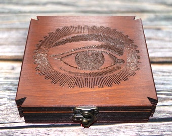 May include: A dark brown wooden box with a detailed engraved design of an eye surrounded by sun rays on the lid. The box has a metal clasp and is made of layered wood. The box is square-shaped.