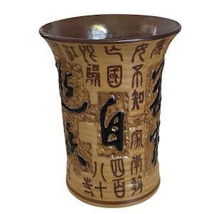 May include: A tan ceramic vase with a flared top and a textured surface. The vase is adorned with black and brown Chinese characters. The interior and rim are a darker brown color.