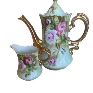 May include: A vintage tea set featuring a teapot and creamer. The set is painted with pink roses and green leaves on a pale green background. The teapot has a gold handle and spout, and the creamer has a matching gold handle.