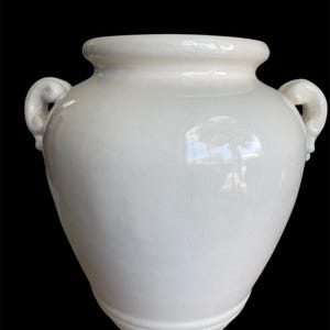 May include: A white ceramic vase with two curved handles and a wide opening. The vase has a rounded body and a slightly textured surface. The vase is set against a black background.