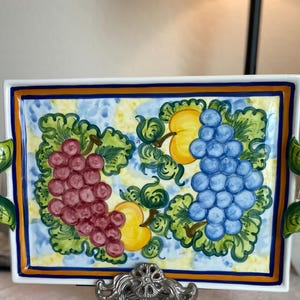 May include: Rectangular ceramic serving tray with a hand-painted design of grapes and peaches. The tray has a white base with a blue and yellow checkered pattern. The border is blue and orange, and the handles are green and orange.