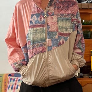 May include: A vintage zip-up jacket in pink, tan, and patterned fabric. The jacket features a high collar, front pockets, and a zipper closure. The patterned section includes blue, pink, and green geometric designs. The jacket is a retro style.