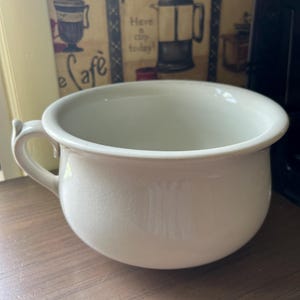 May include: A white ceramic chamber pot with a rounded body, a wide opening, and a single side handle. The surface features a subtle, fine crackle glaze pattern throughout the exterior, giving it a textured appearance while maintaining a smooth finish.