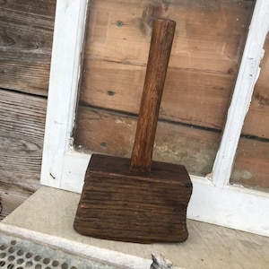 May include: A dark brown wooden mallet with a long handle. The mallet is sitting on a light gray surface.