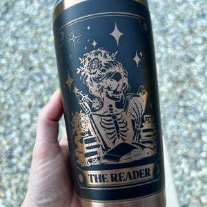 May include: Black and copper-colored insulated tumbler featuring a tarot card design. The design includes a skeleton figure with floral accents, reading a book, with the words "THE READER" at the bottom. The tumbler has a copper-colored rim and base.