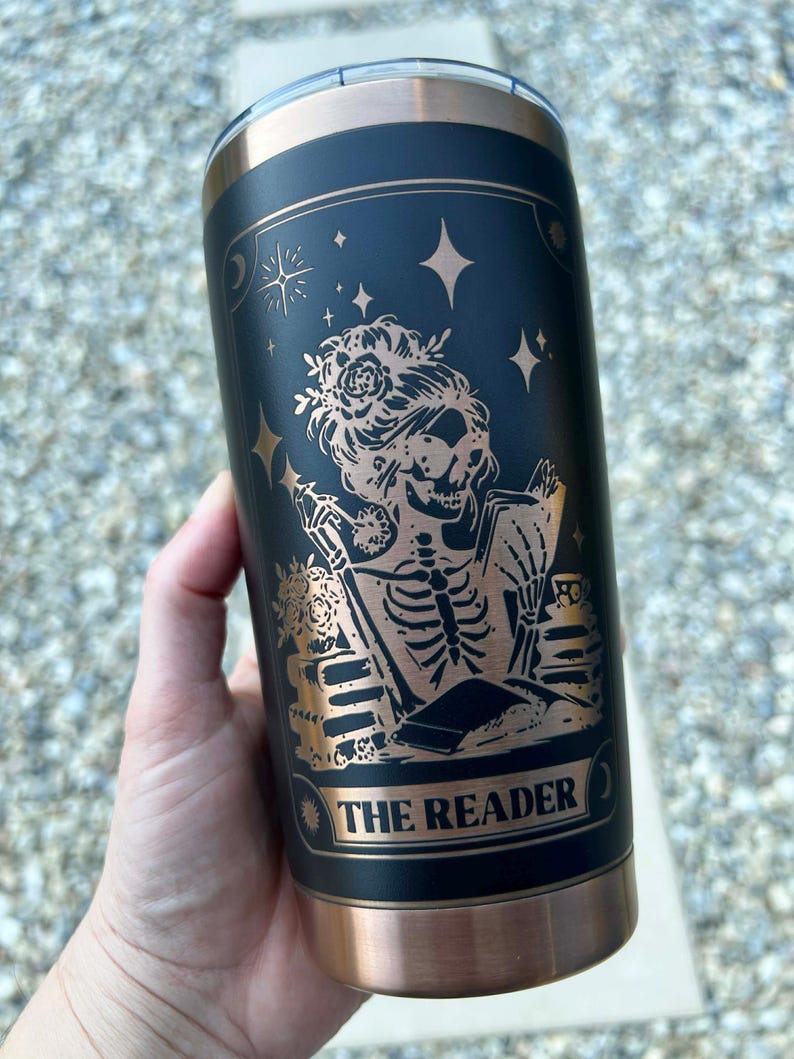 May include: Black and copper-colored insulated tumbler featuring a tarot card design. The design includes a skeleton figure with floral accents, reading a book, with the words "THE READER" at the bottom. The tumbler has a copper-colored rim and base.