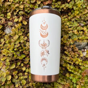 Puede incluir: Un vaso de acero inoxidable blanco con tapa y base de color cobre. El vaso presenta un dise&ntilde;o de cinco lunas en diferentes fases con estrellas y detalles florales en l&aacute;mina de cobre.