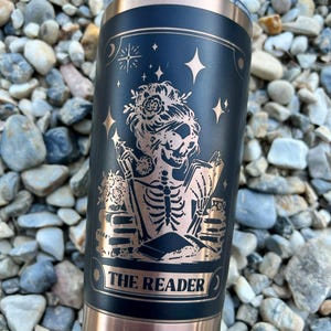 May include: Black and copper-colored insulated tumbler with a design featuring a skeleton reading a book, adorned with floral elements and stars. The text "THE READER" is displayed on the tumbler.