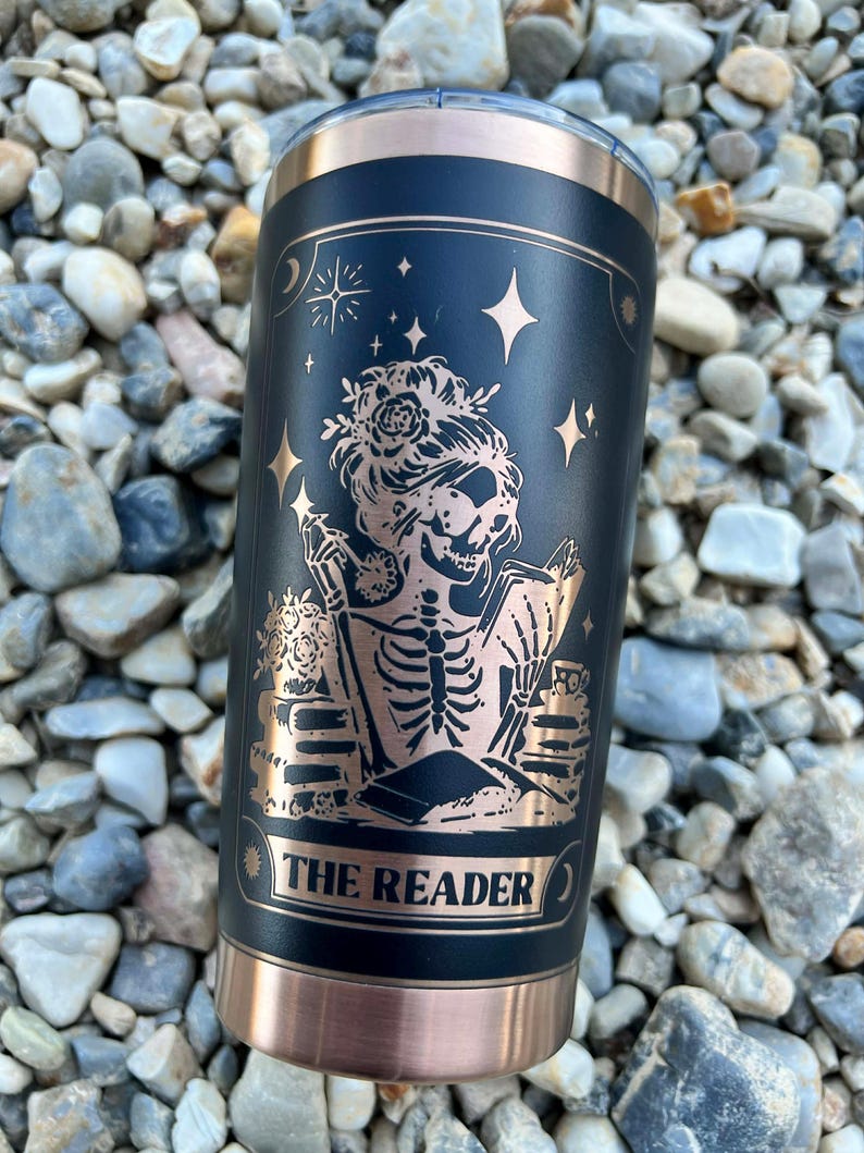 May include: Black and copper-colored insulated tumbler with a design featuring a skeleton reading a book, adorned with floral elements and stars. The text "THE READER" is displayed on the tumbler.