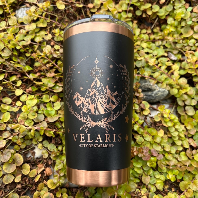 Puede incluir: Un vaso de acero inoxidable negro y cobre con un dise&ntilde;o de una cordillera, estrellas y un sol en una corona. El texto "VELARIS - CITY OF STARLIGHT" est&aacute; impreso en el vaso.