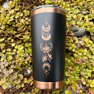 Puede incluir: Un vaso de acero inoxidable negro y cobre con un dise&ntilde;o de lunas y estrellas en color cobre. El vaso est&aacute; sobre un lecho de follaje verde.