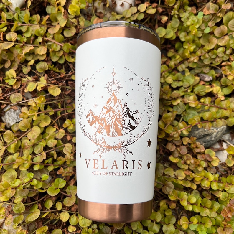 Puede incluir: Un vaso de acero inoxidable blanco con tapa de cobre. El vaso presenta un dise&ntilde;o dorado de una cordillera con un sol y estrellas, y el texto "VELARIS - CITY OF STARLIGHT."