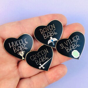 May include: Four black heart-shaped enamel pins with gold accents. Each pin features a different Hogwarts house crest and the text "[House Name] BABE".