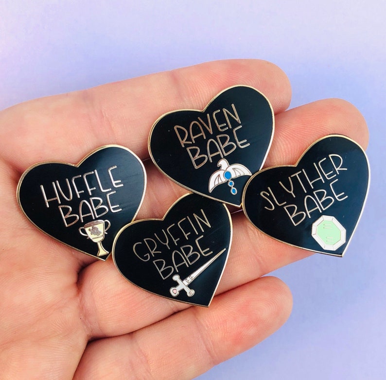 May include: Four black heart-shaped enamel pins with gold accents. Each pin features a different Hogwarts house crest and the text "[House Name] BABE".