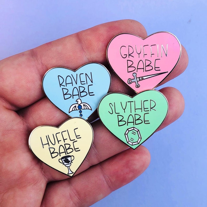 May include: Four heart-shaped enamel pins with different Hogwarts house colors and crests. The pins read "Gryffin Babe", "Raven Babe", "Slyther Babe", and "Huffle Babe".